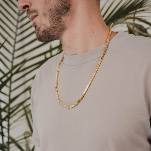 A close-up lifestyle shot of a man wearing a gold byzantine chain necklace over a light gray t-shirt, with a blurred background of green palm leaves.